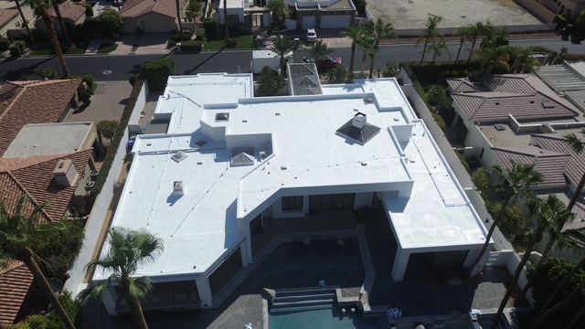 Aerial view of a modern, white-roofed house with a pool and surrounding palm trees. The building is in a residential neighborhood.