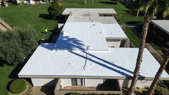 Aerial view of a white flat roof on a building, with a palm tree casting a shadow. A person is visible near the edge.