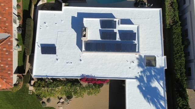 Aerial view of a white flat roof with solar panels. The roof is on a residential building, surrounded by greenery and adjacent buildings.
