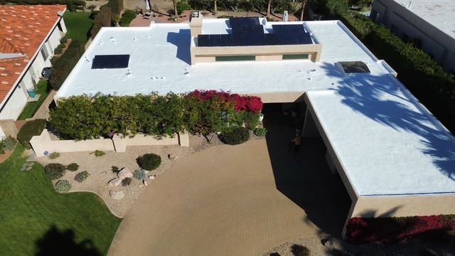 Aerial view of a modern house with a white flat roof, solar panels, and a long driveway lined with landscaping.