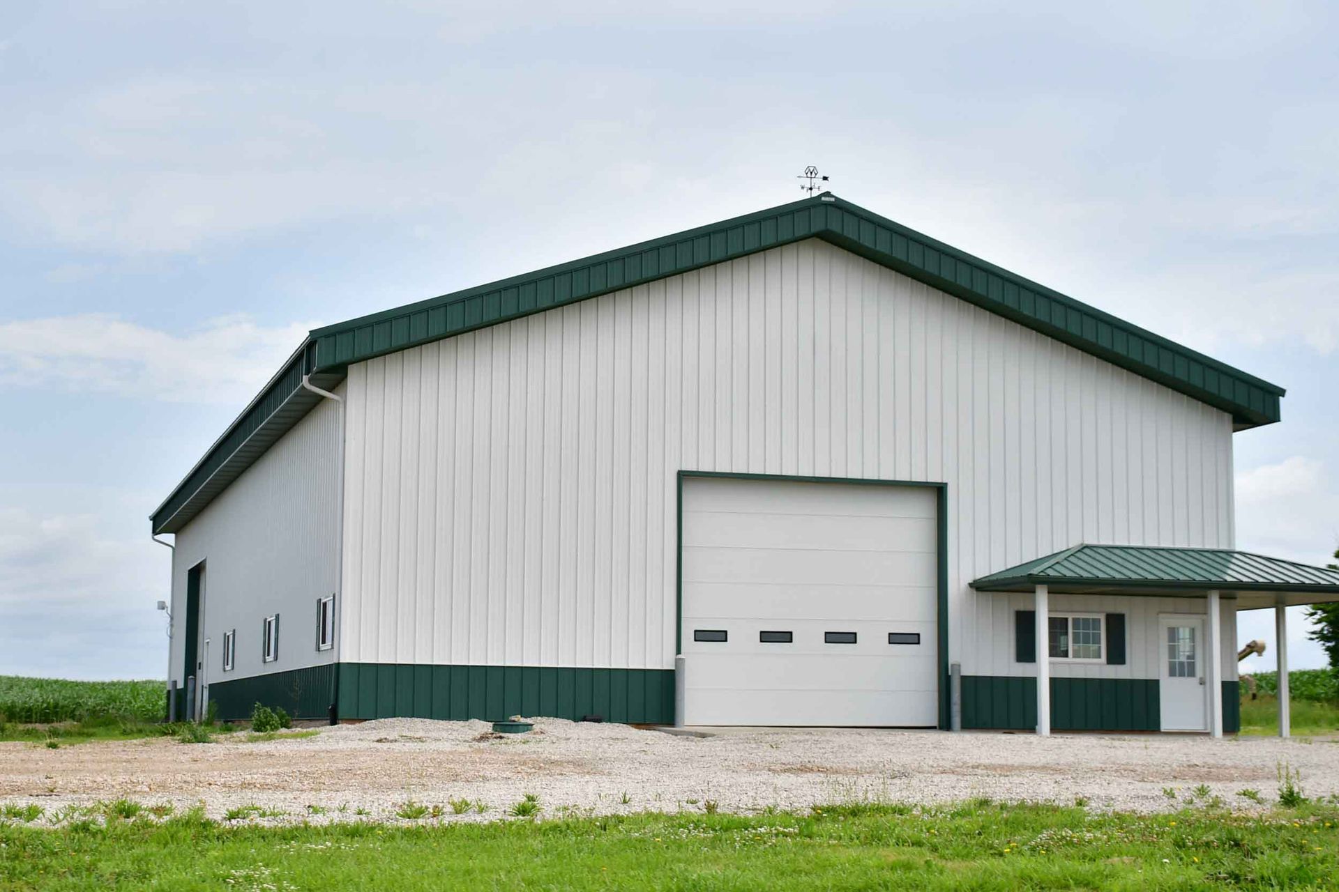 White and green metal barn with a large garage door, gravel ground, and small office.