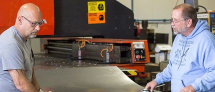 Two men are standing next to each other in front of a Turret Punch machine in a factory.