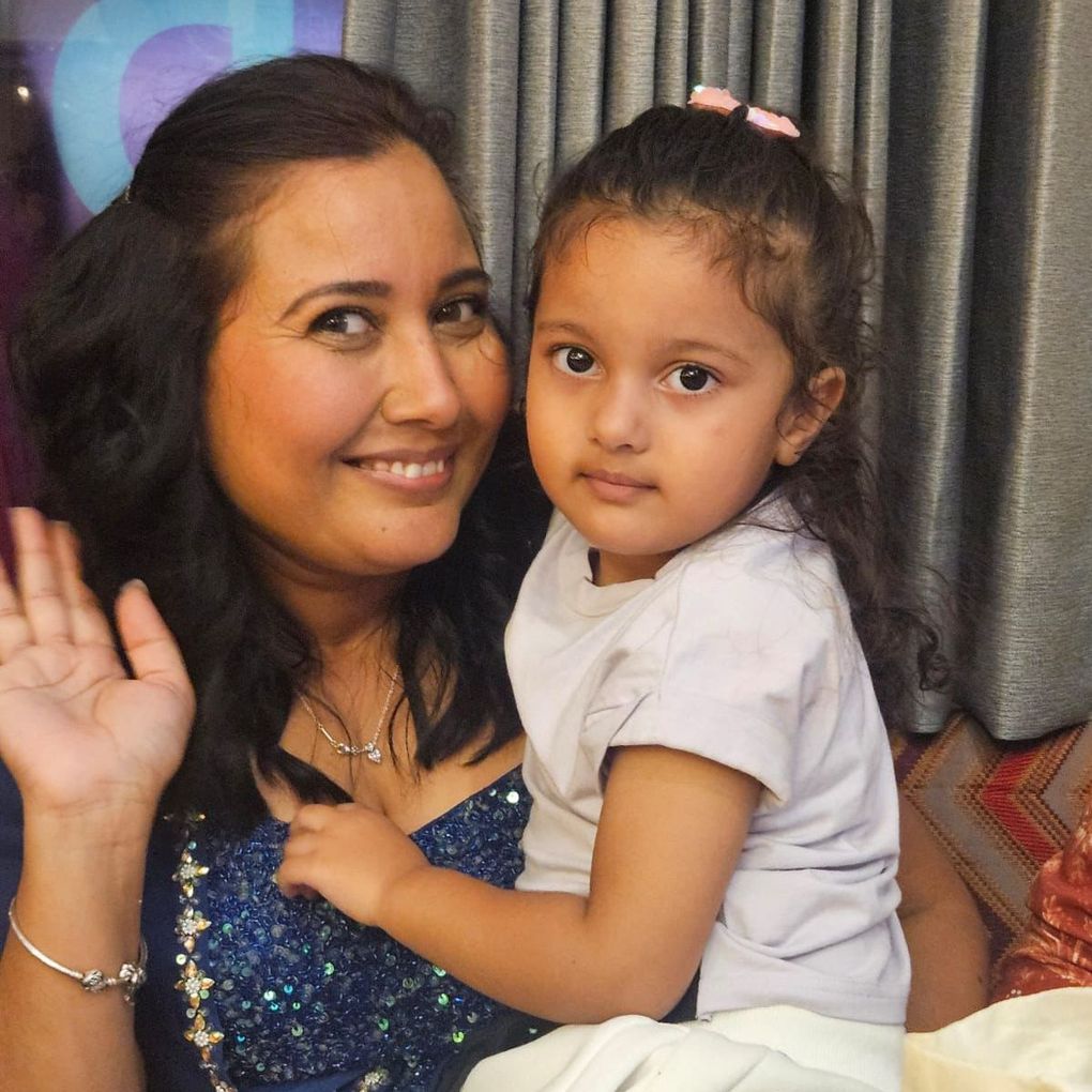 Woman with dark hair waves; holds a young child in a light shirt; indoor setting.