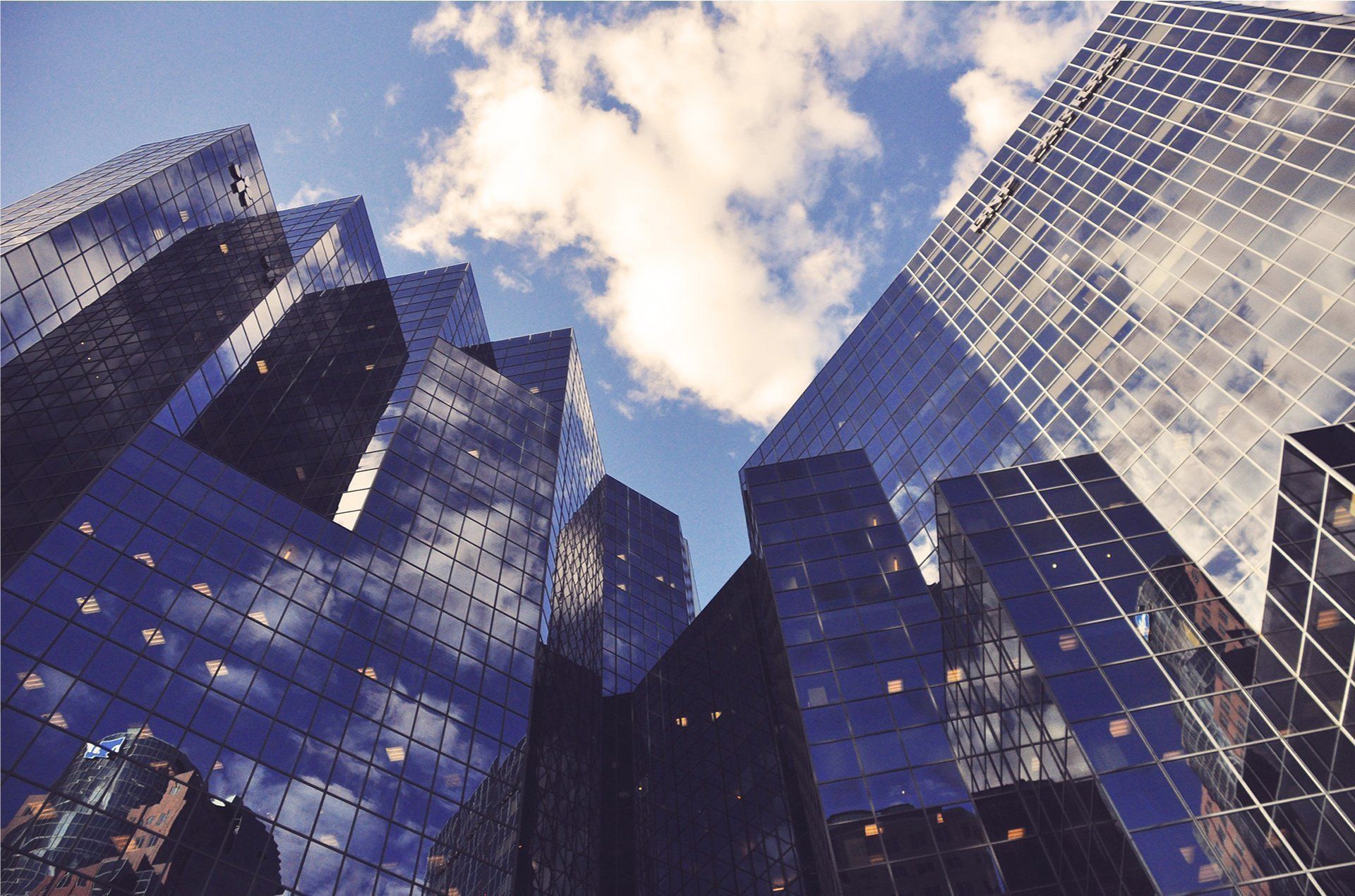 Skyscrapers with mirrored glass reflect clouds and blue sky, viewed from a low angle.