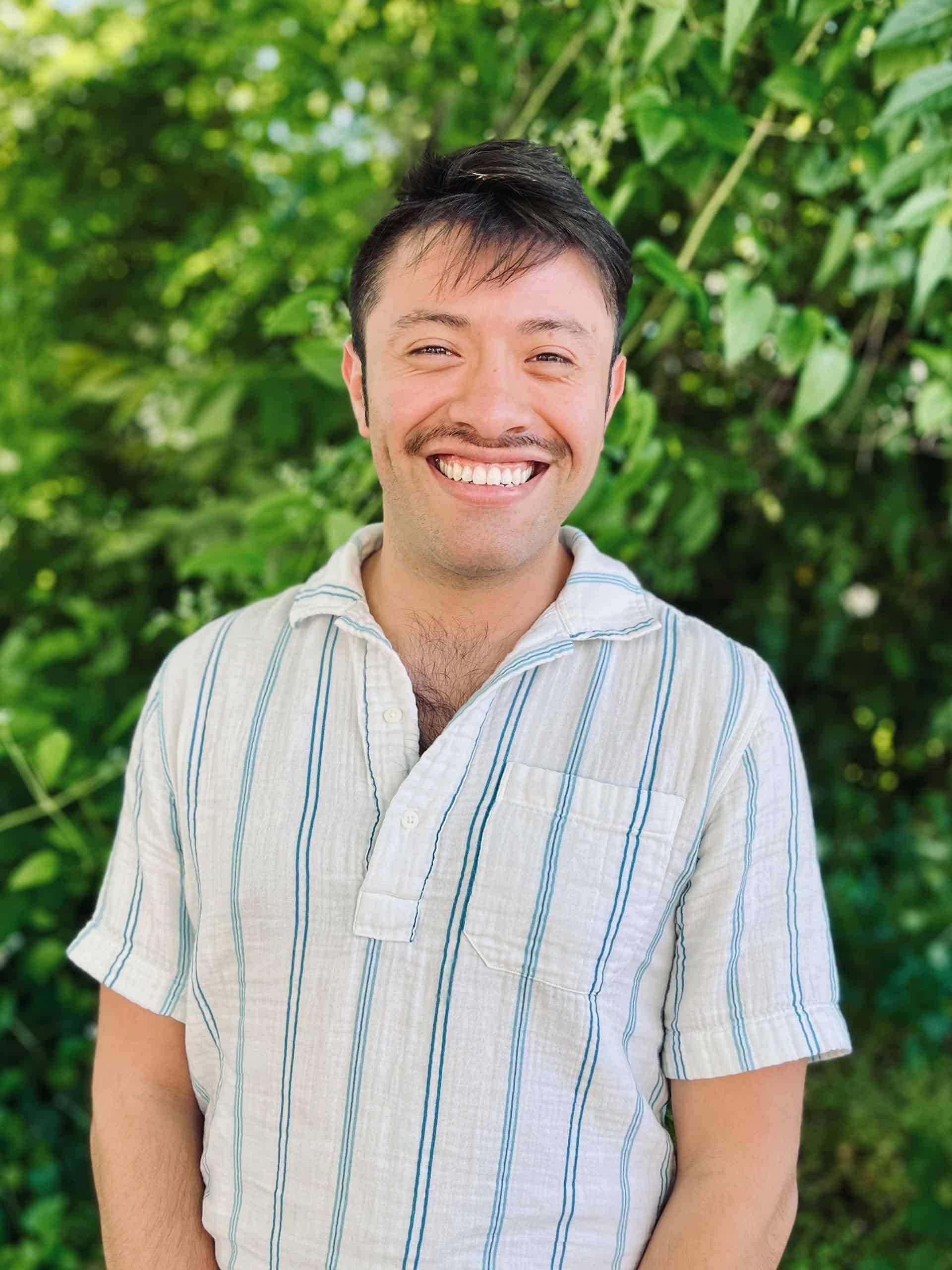 A man in a striped shirt is smiling in front of a tree.