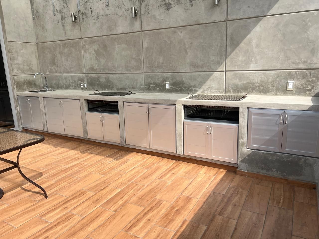 Cocina al aire libre con gabinetes y electrodomésticos de acero inoxidable contra una pared de concreto, piso de madera.