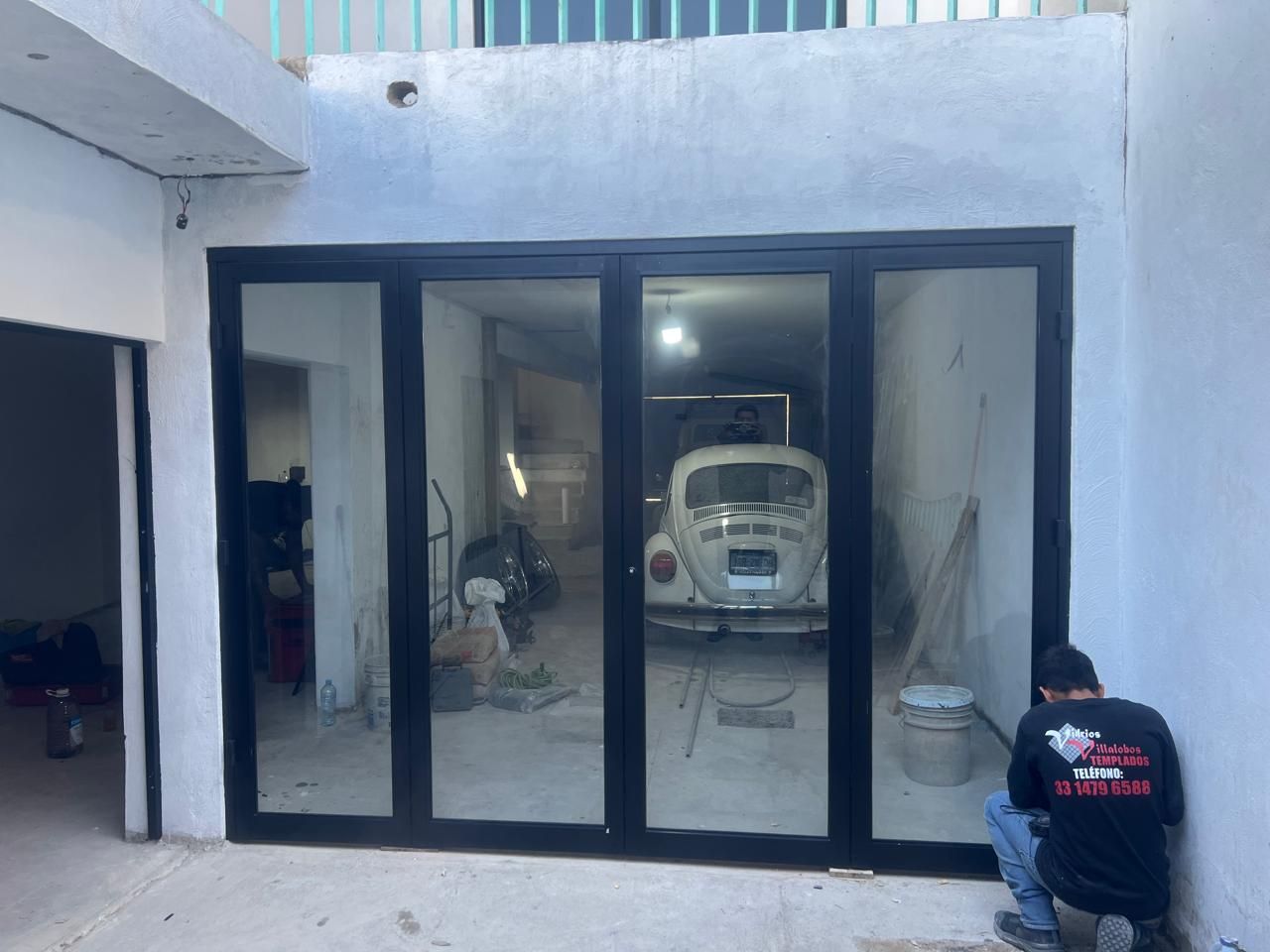 Unas puertas de cristal con marco negro dan a un garaje con un coche blanco dentro. Una persona trabaja a la derecha.