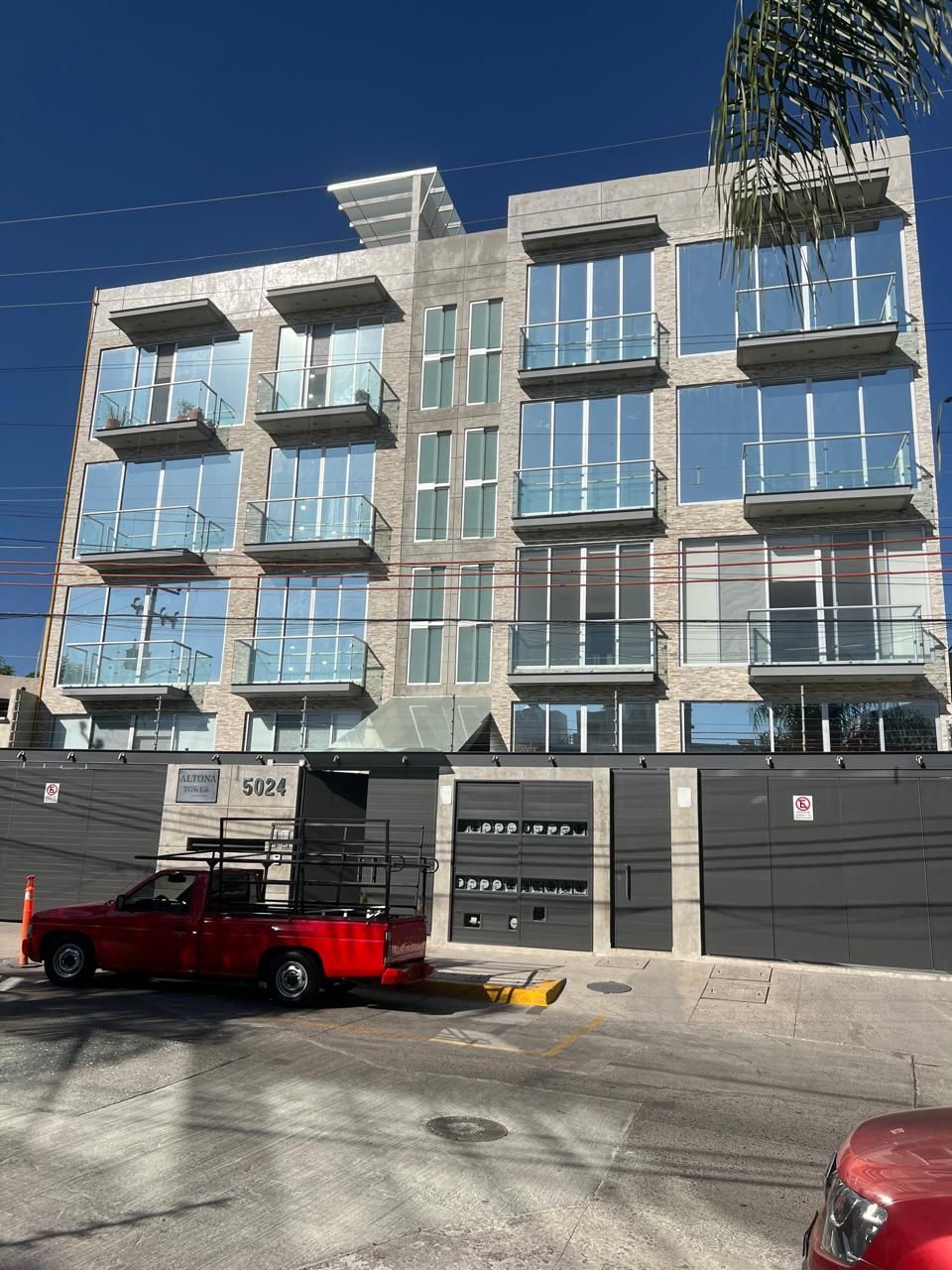 Edificio de departamentos moderno con amplios ventanales y balcones, camioneta roja estacionada en la calle.