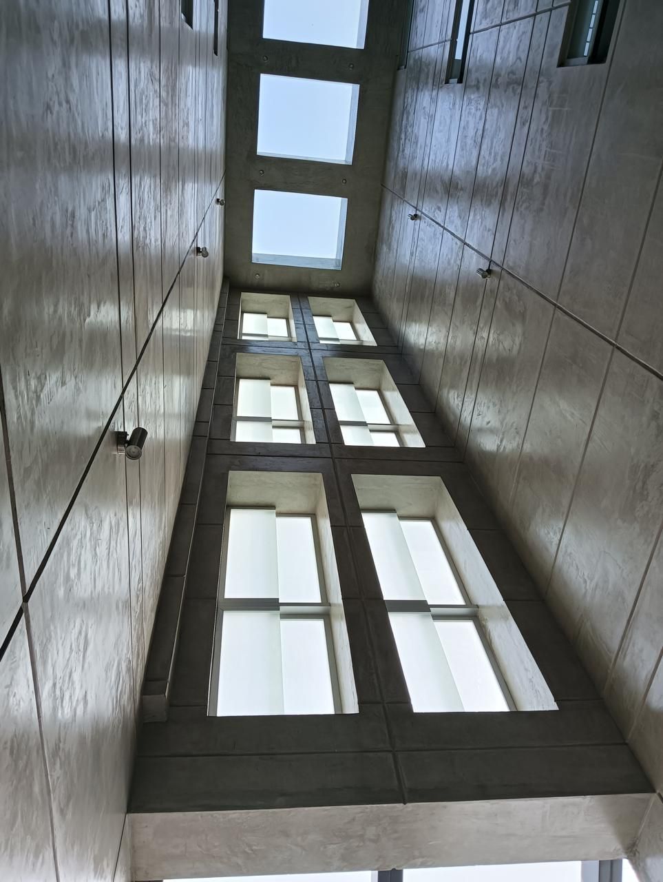 Espacio arquitectónico interior estrecho con ventanas altas y tragaluces, visto desde abajo.