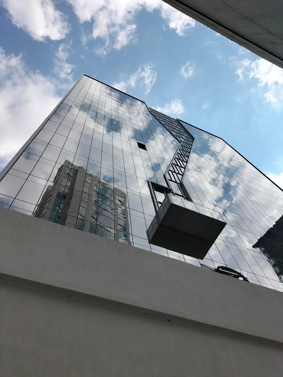 Rascacielos de cristal que refleja el cielo y otros edificios. Una plataforma gris cuelga de él.