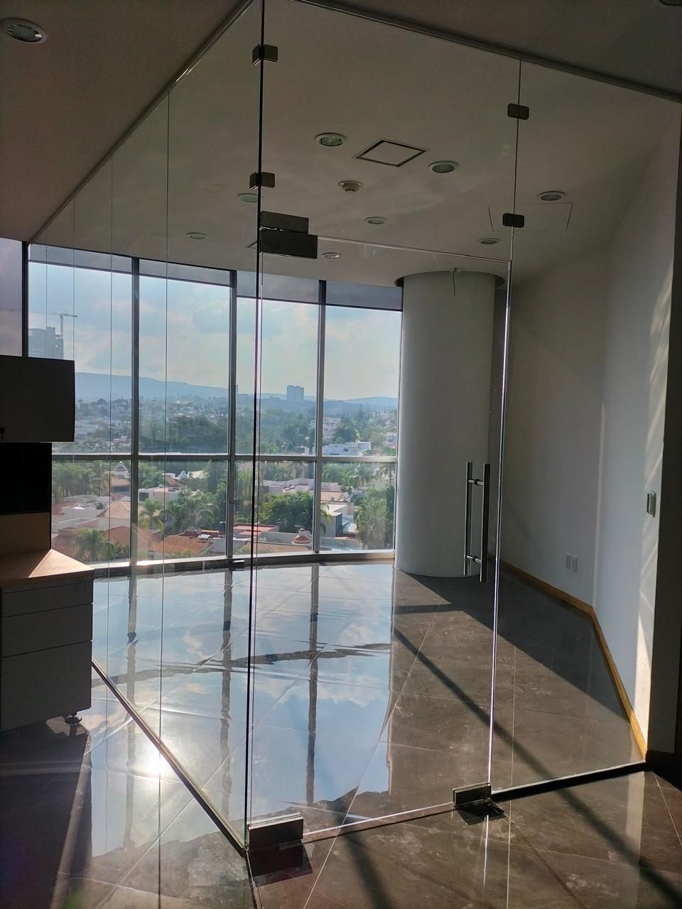 Interior de oficina con paredes de vidrio, vista a la ciudad y luz natural reflejada en un piso brillante.