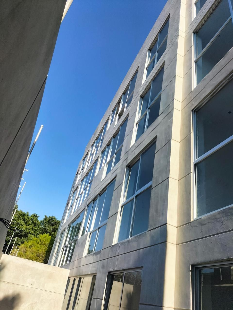 Edificio gris de varios pisos con muchas ventanas y un cielo azul brillante encima.