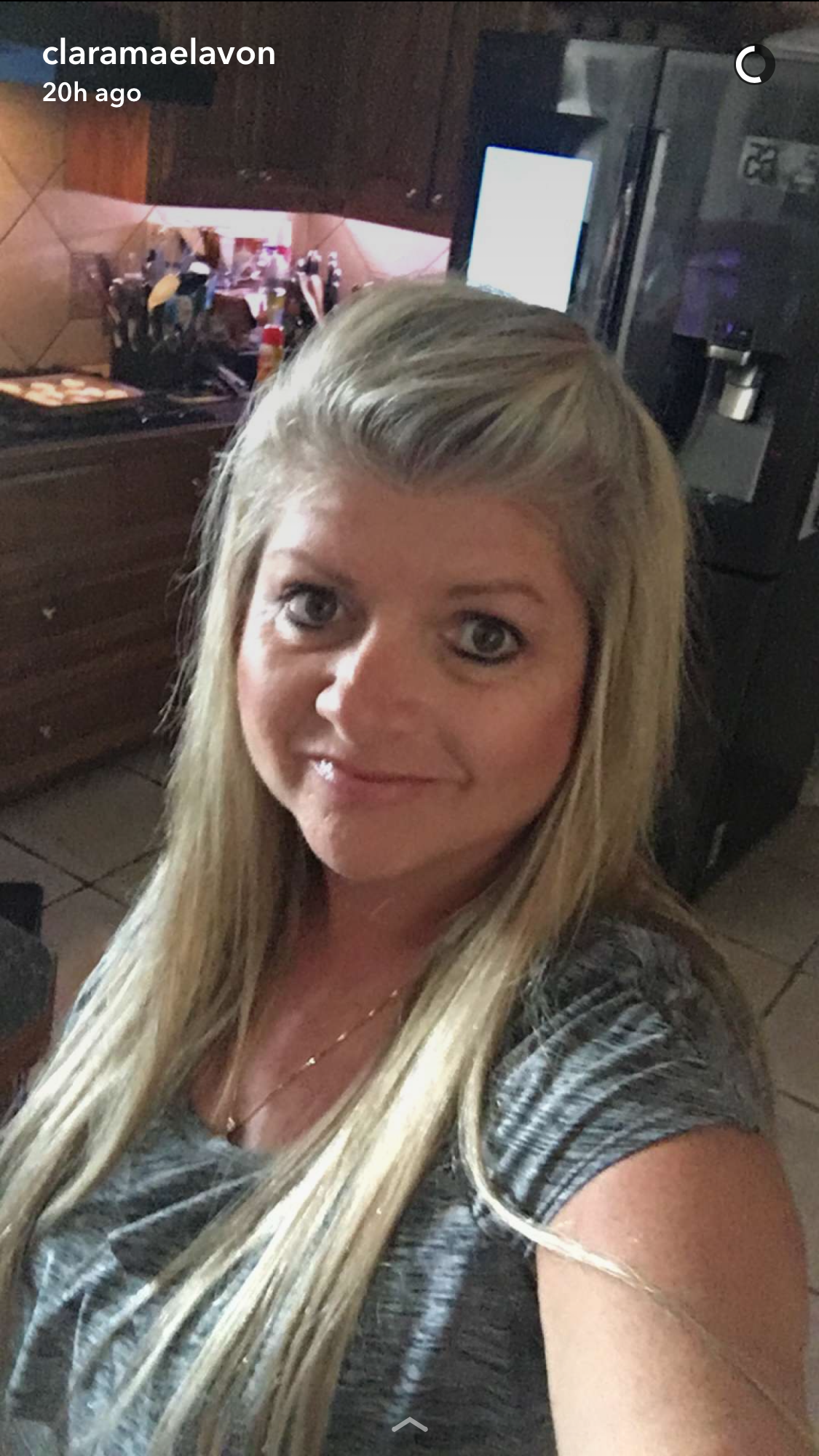 Blond woman in a gray shirt smiles. Kitchen background with appliances visible.