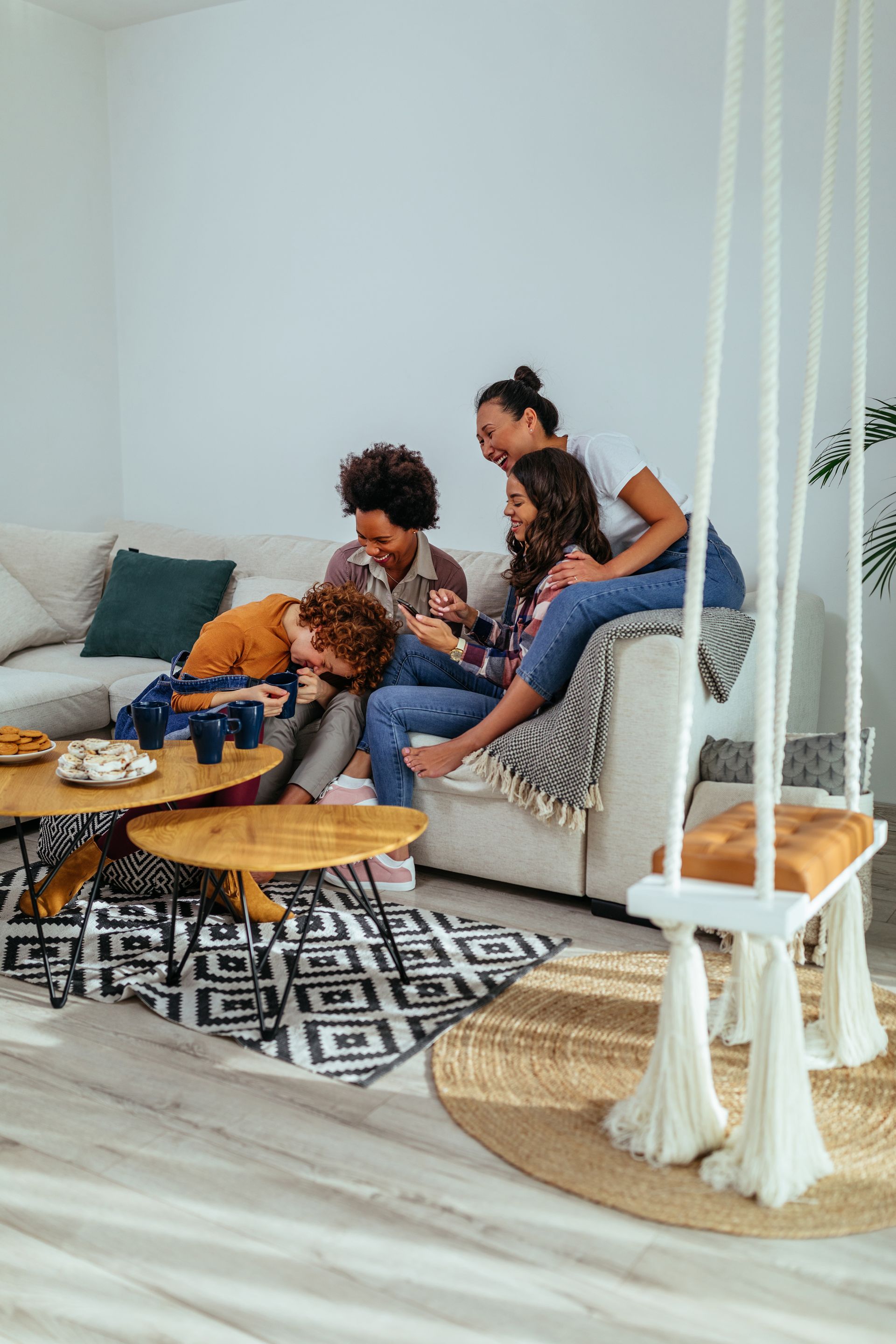 Four people laughing on a couch, near a swing and coffee tables with snacks, in a living room.