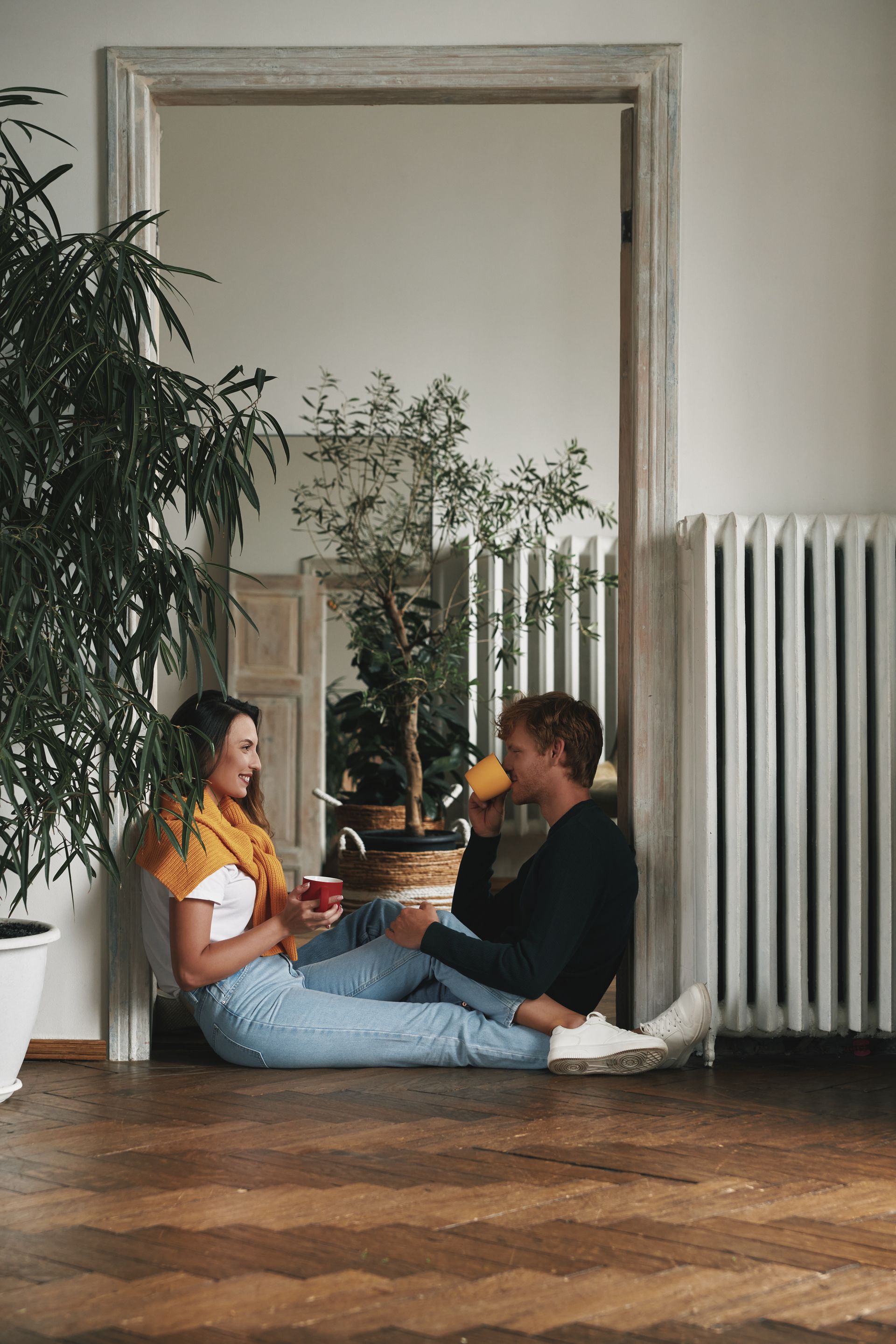 Couple sitting in doorway, drinking from mugs, smiling. Indoor, plants, radiator, wood floor.