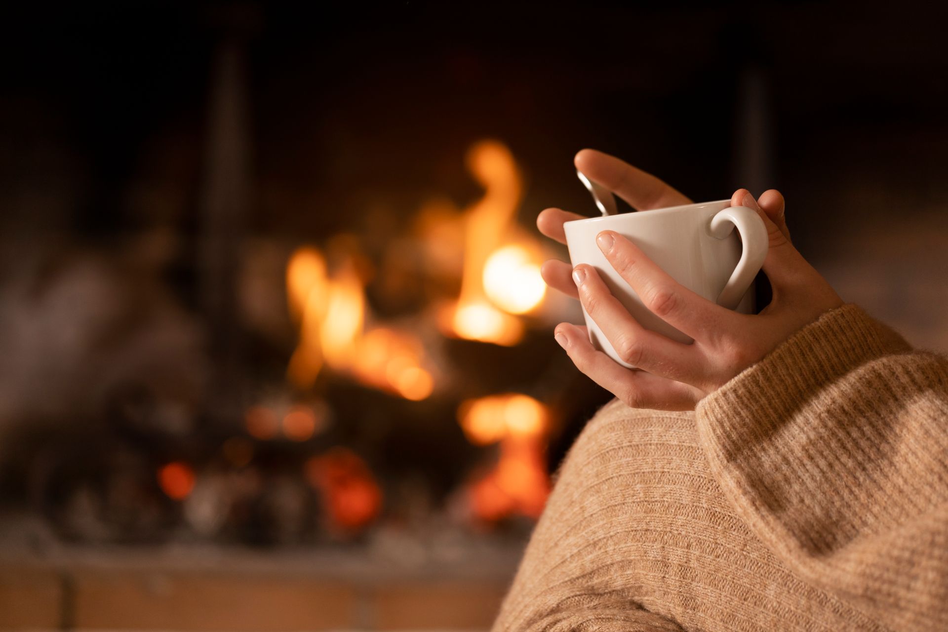 Hands holding a white mug near a fireplace, person wrapped in a beige sweater, flames in the background.