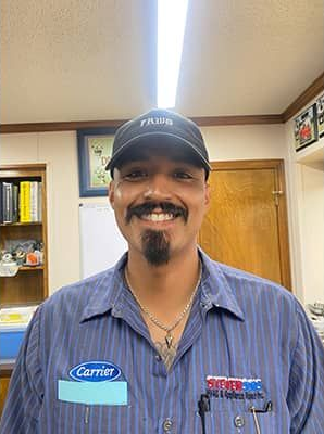 Man in blue work shirt and cap, smiling. Inside a workshop with shelving and a door.