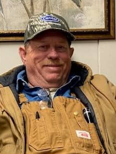 Man in camo hat, overalls, and jacket, smiling.