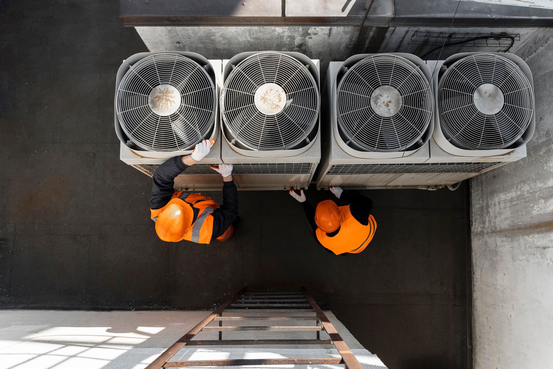 Two workers in safety vests inspect HVAC units on a rooftop, with a ladder.