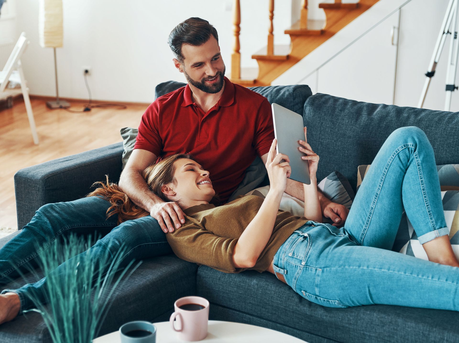Couple relaxing on a couch, looking at a tablet, smiling. Cozy living room setting.