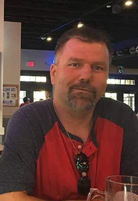 Man in red and navy shirt with a short beard smiles, seated in a restaurant.