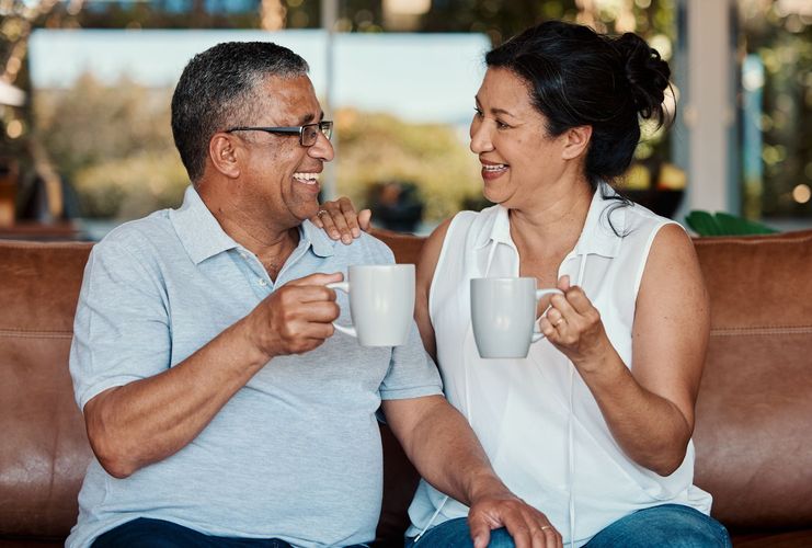 Couple on a brown couch smiling and holding mugs. Man has his arm around woman's shoulder.