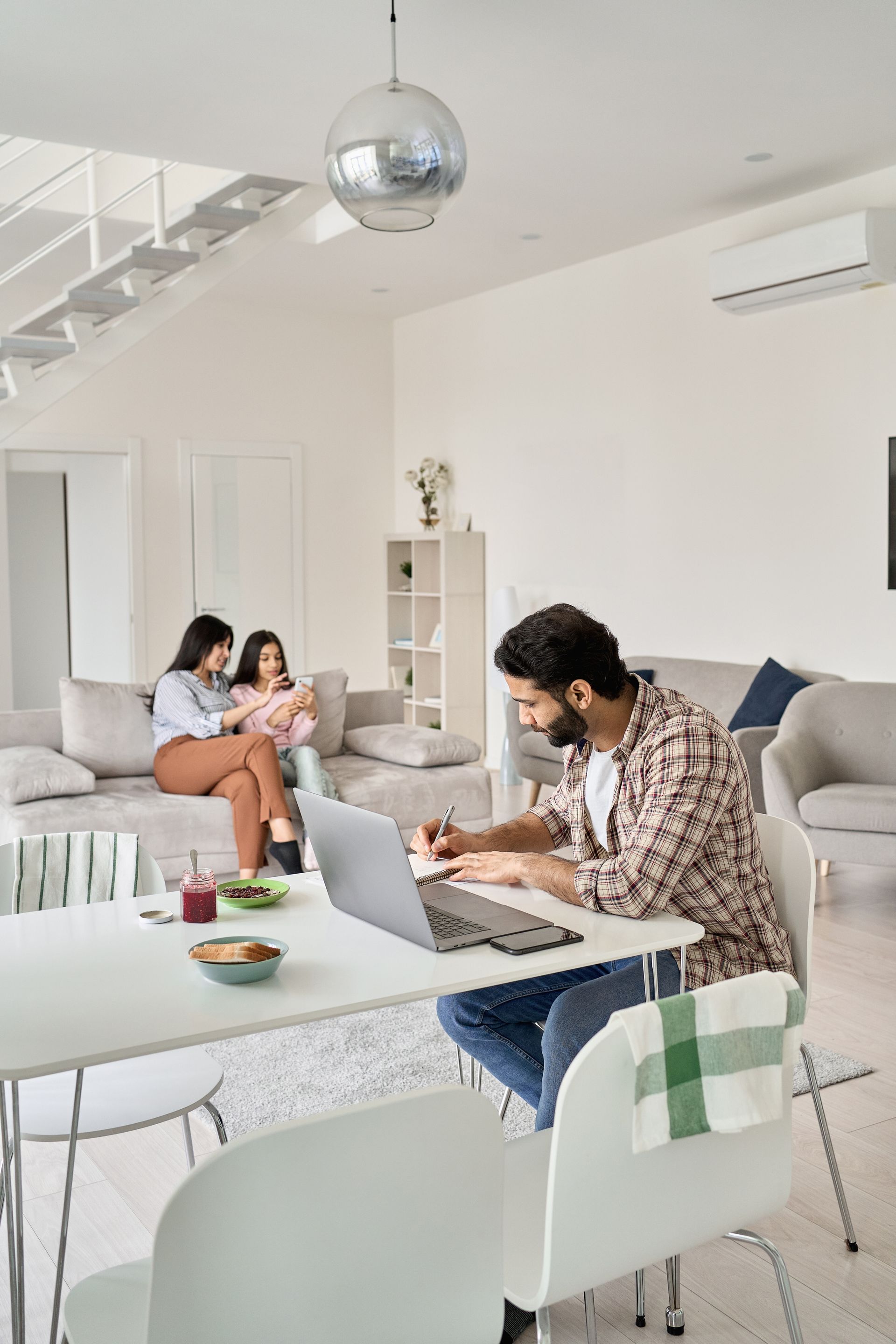 Man working on laptop at table, women on sofa in bright living room.