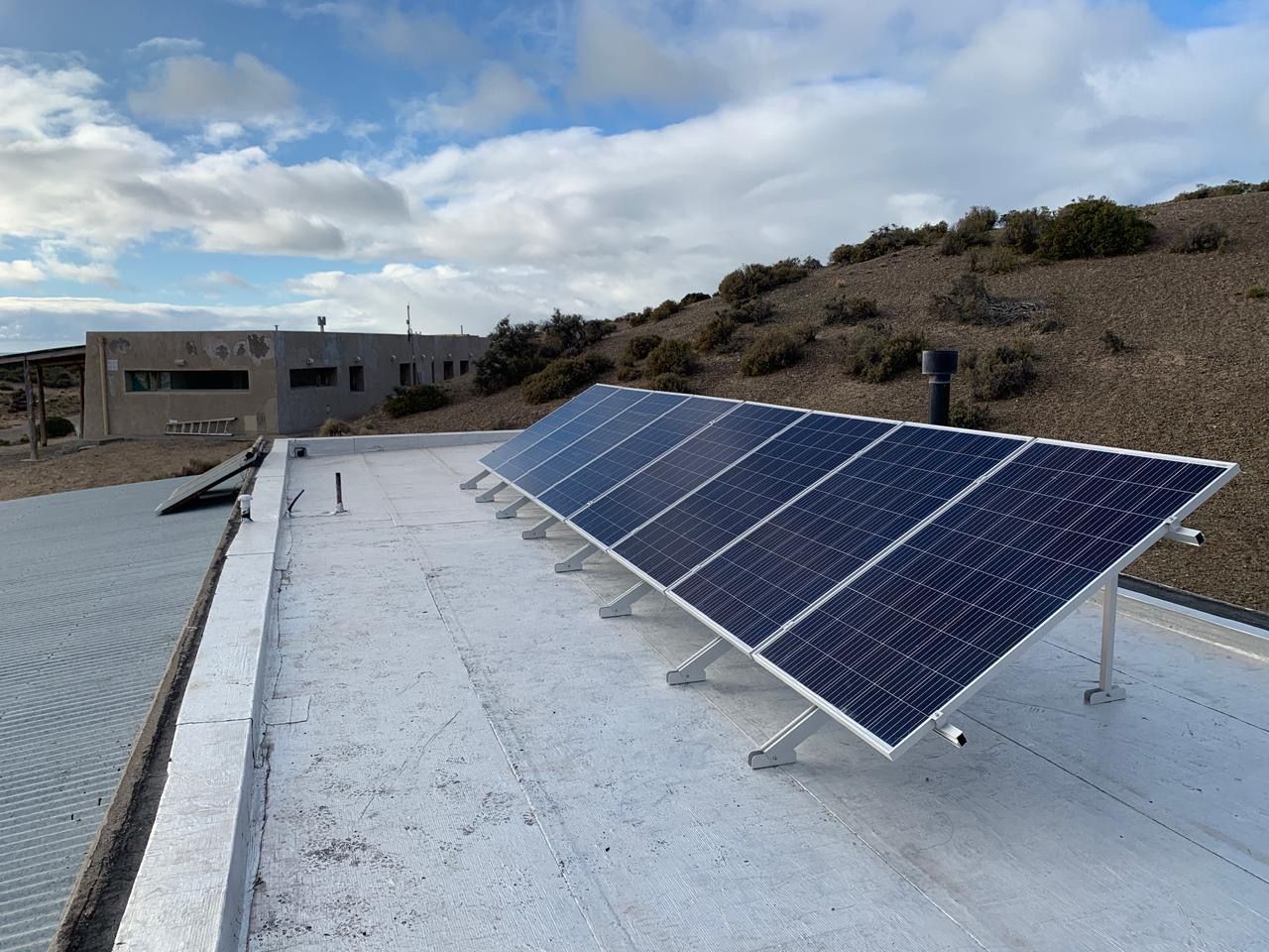 Paneles solares sobre un tejado plano, orientados hacia el cielo, con un edificio y una ladera al fondo.