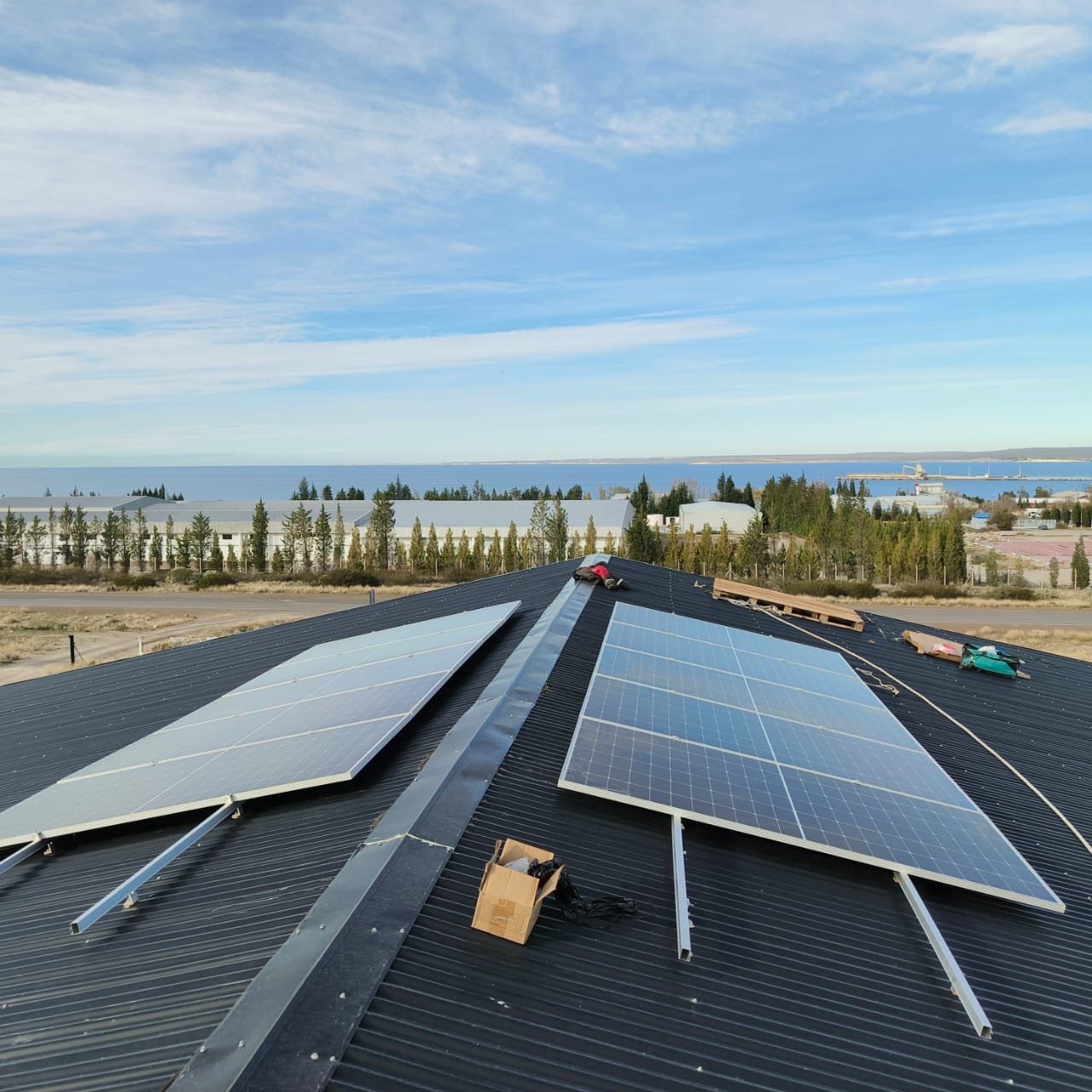 Paneles solares sobre un techo oscuro con una vista panorámica del agua y los árboles bajo un cielo azul.