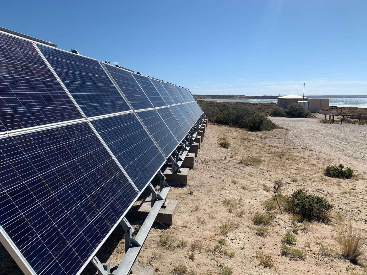 Paneles solares sobre una estructura de metal, en un entorno desértico con un edificio y agua a lo lejos.