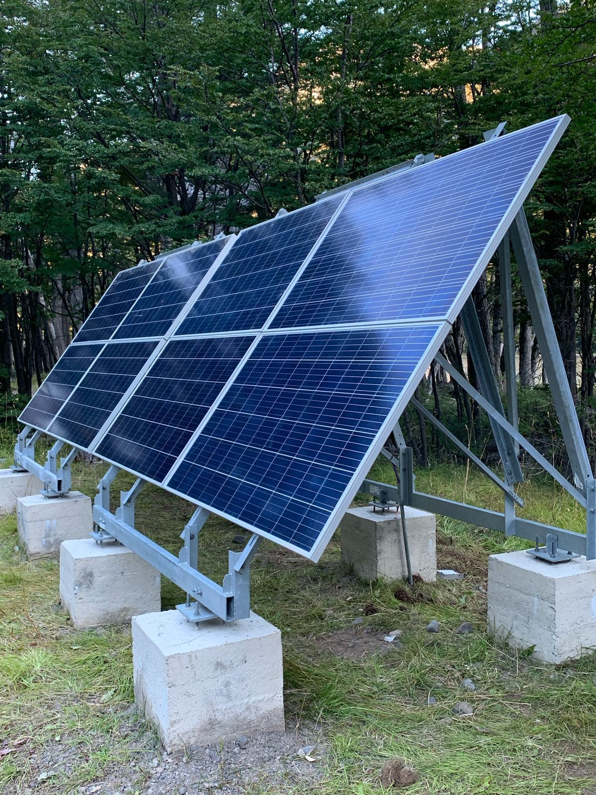 Paneles solares montados sobre un marco de metal, colocados sobre bloques de hormigón en una zona verde con árboles al fondo.