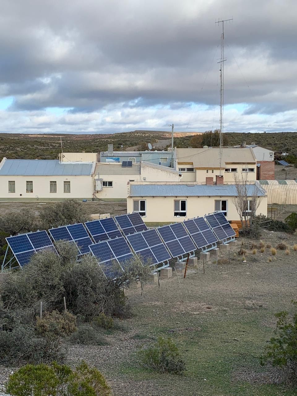 Paneles solares frente a un edificio con una antena alta. Cielo nublado, paisaje natural.