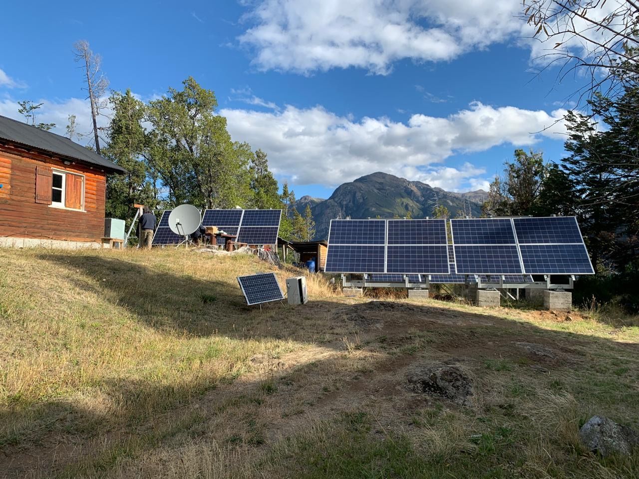 Paneles solares en un patio con césped, con un edificio de ladrillo y montañas al fondo bajo un cielo azul.