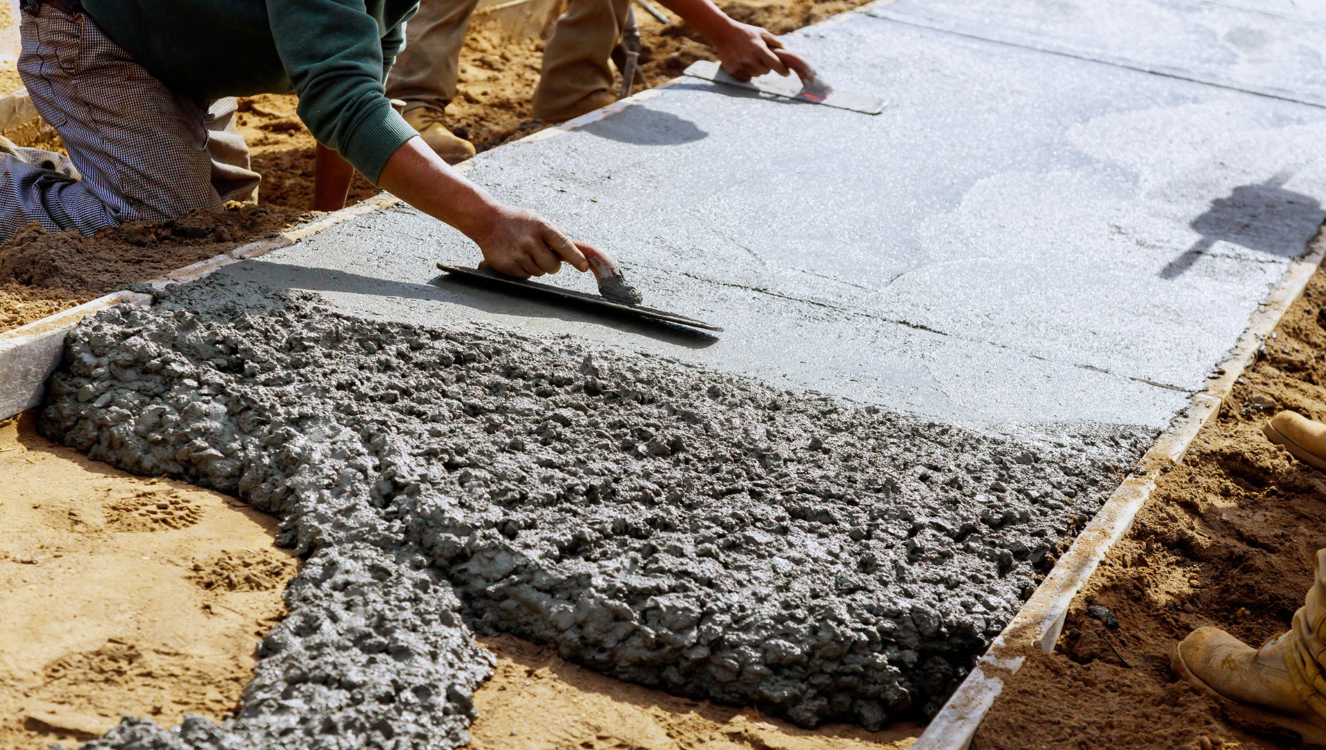 Workers smoothing wet concrete with trowels in a wooden form on a dirt surface.