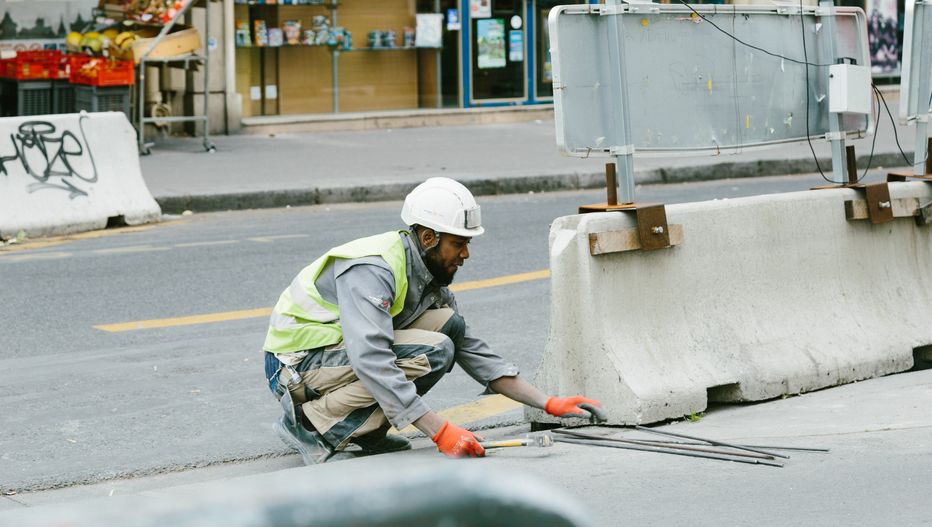 Construction worker in safety vest and helmet crouches to work on pavement in street, near barriers.