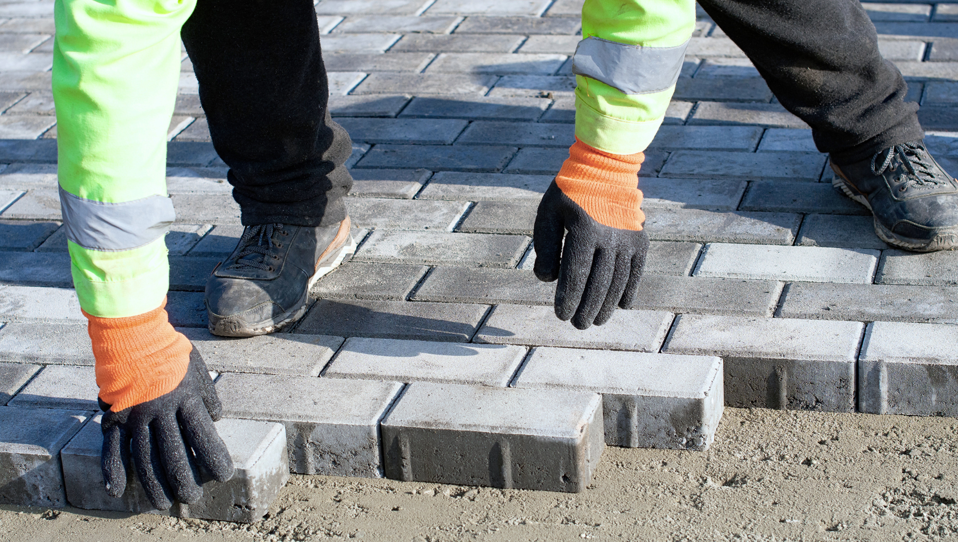 Construction worker placing paving stones, wearing orange gloves and a reflective vest.