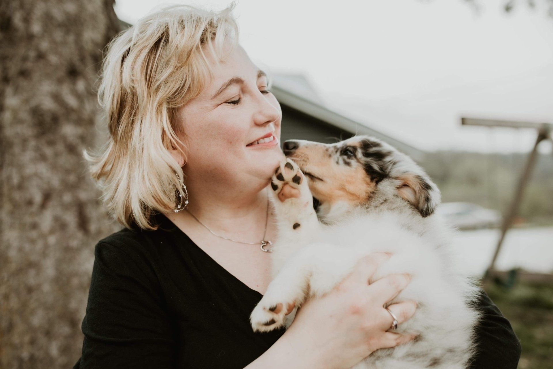 Woman smiling, holding a puppy, both looking content. Puppy's paw on woman's face. Outdoors, overcast day.