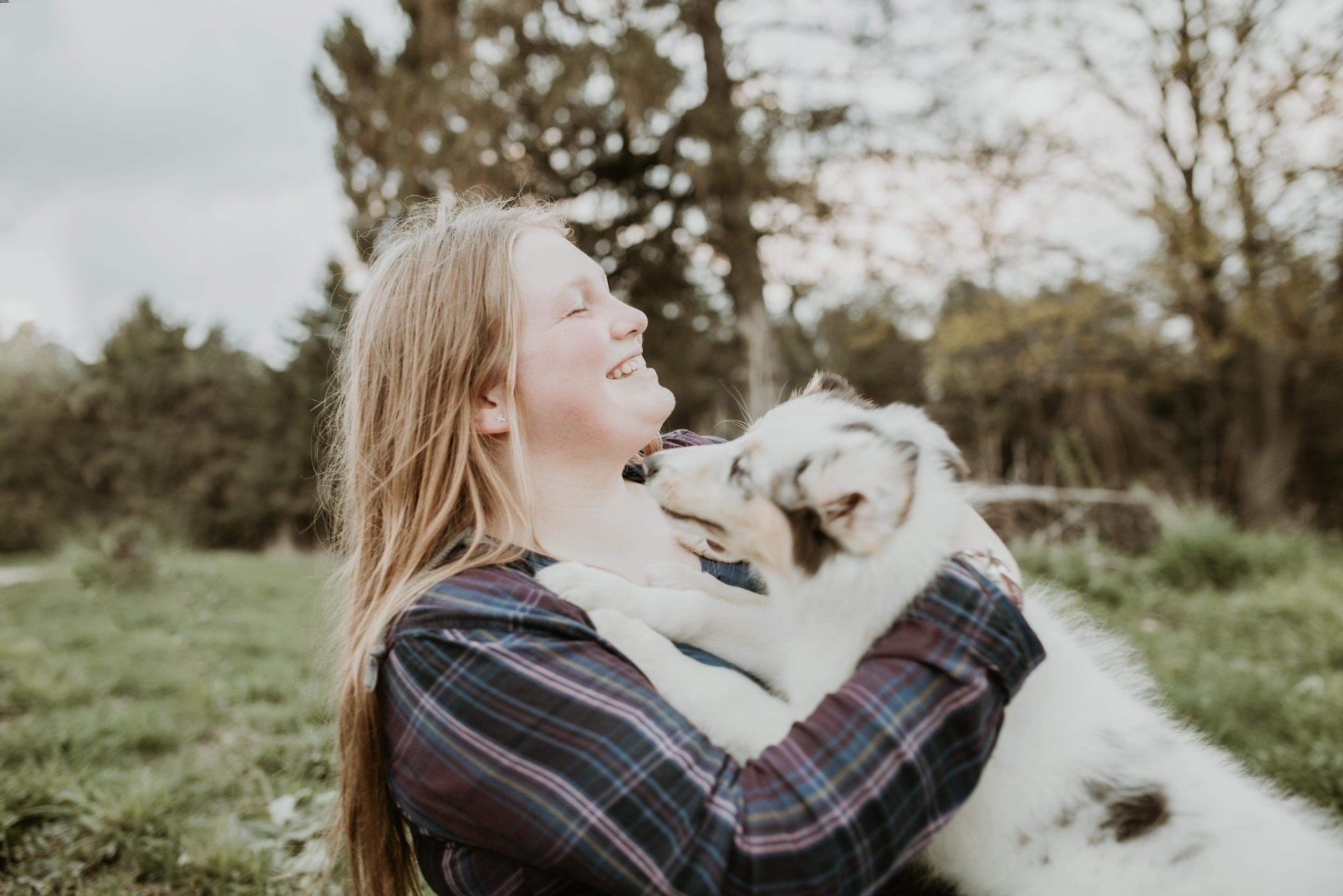 Woman laughing, hugging a blue merle dog outdoors on a grassy lawn.