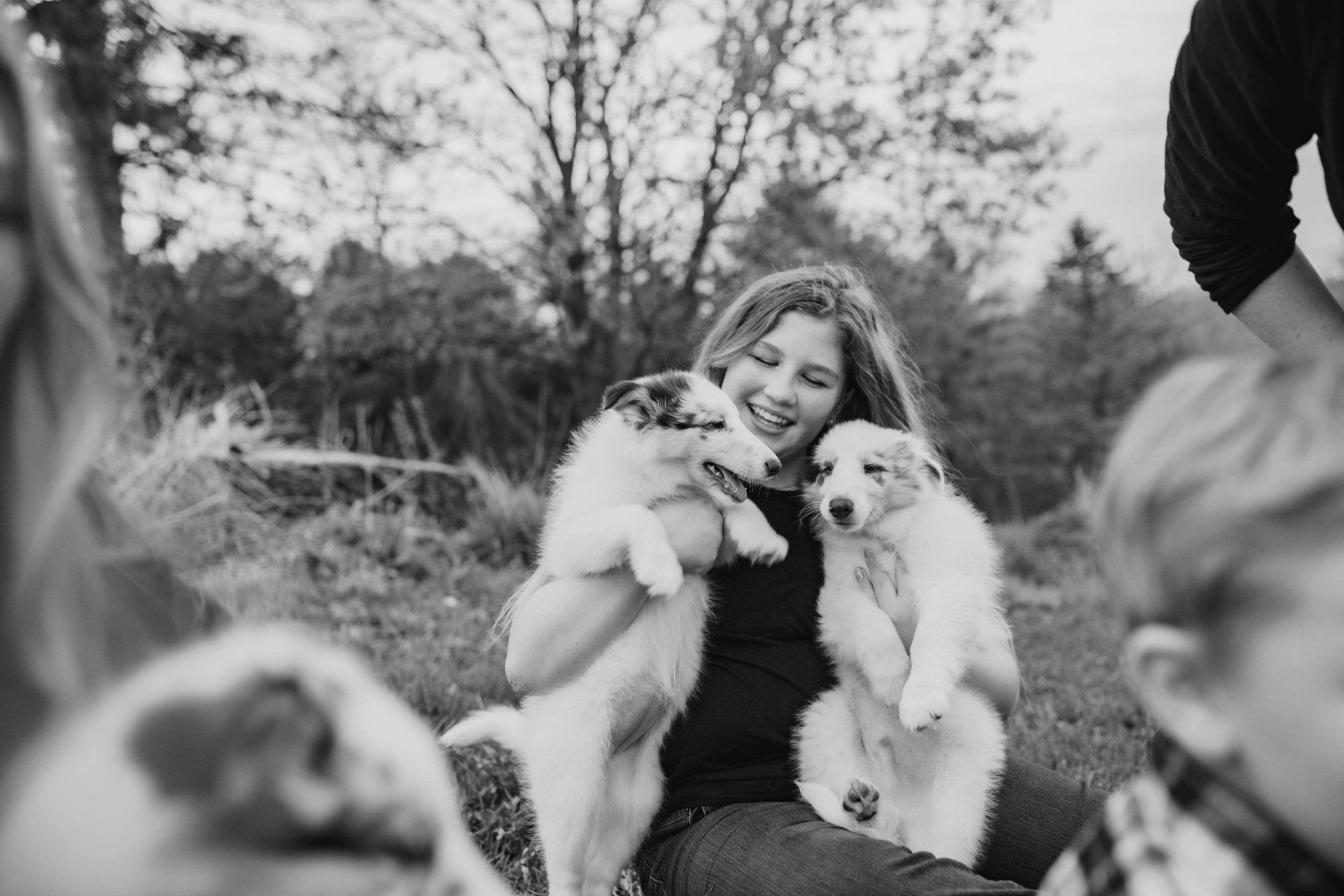 Woman smiling, holding two puppies. Outdoors, other people and dogs in background. Black and white.