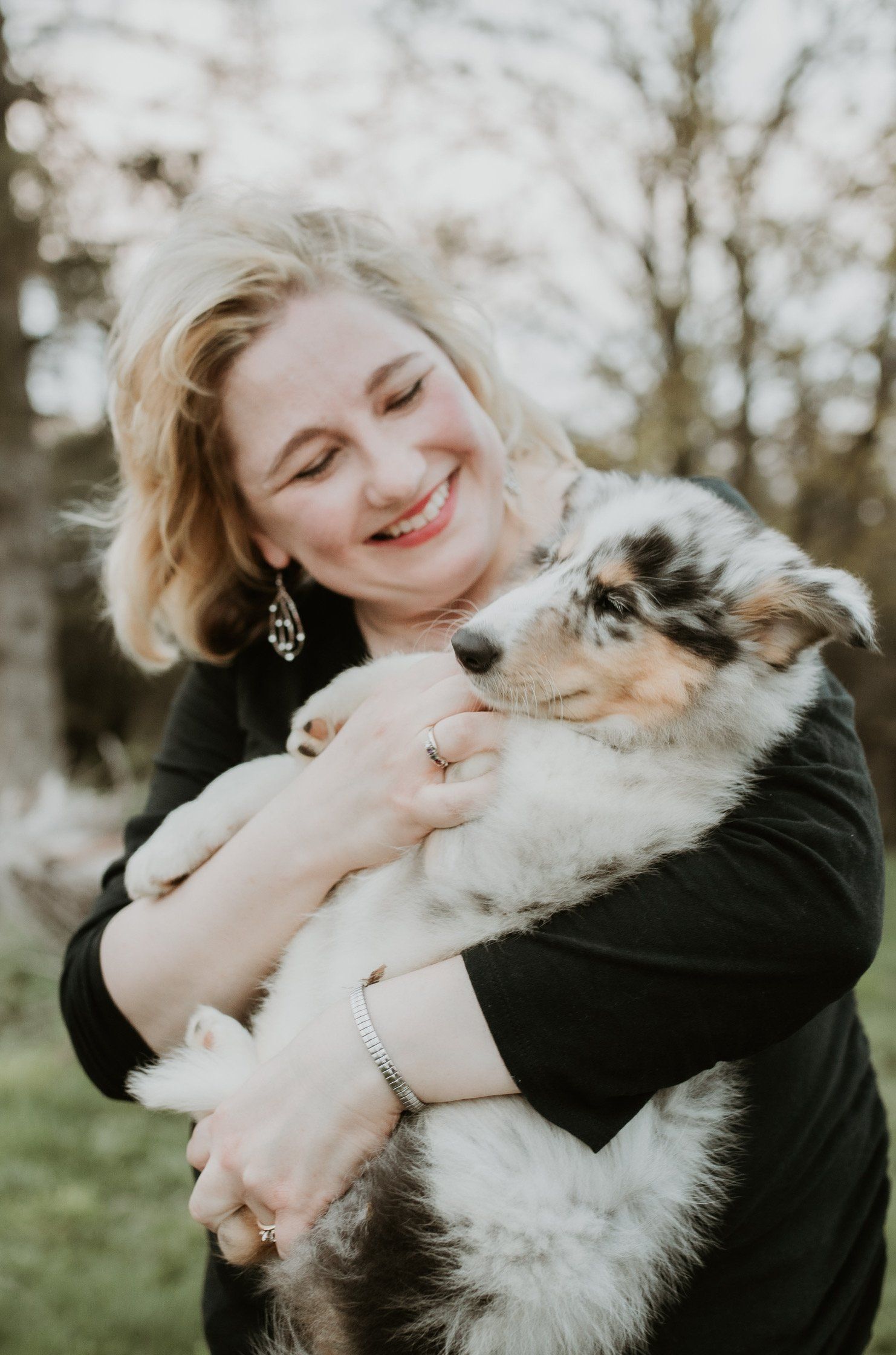 Woman with blond hair smiles, holding a fluffy blue merle puppy outdoors.