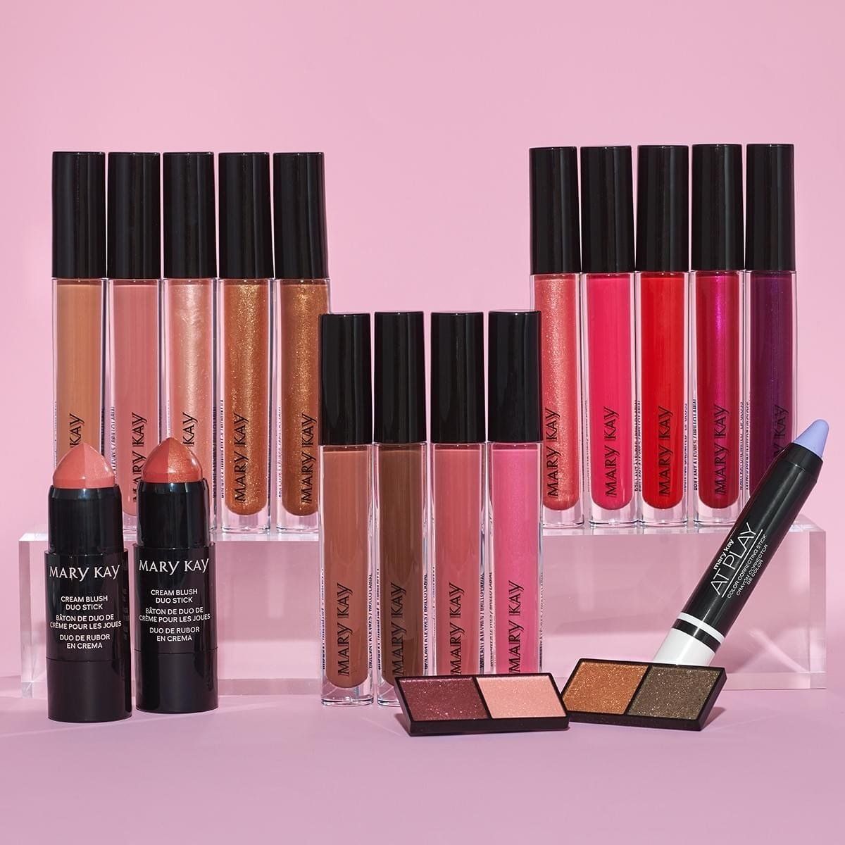 Cosmetics set: lip glosses, blush sticks, eyeshadows, and eye pencil on pink surface.