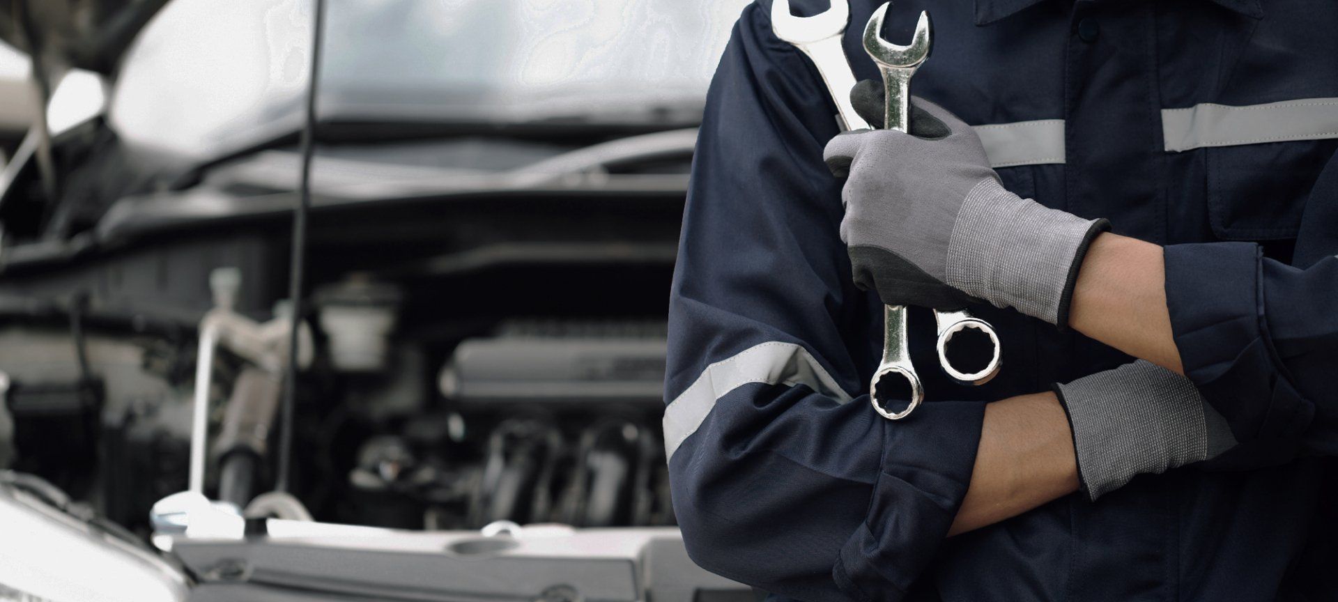 A mechanic in a blue jumpsuit holding wrenches, working on a car with the hood open. | Bencomo Motorsport LLC
