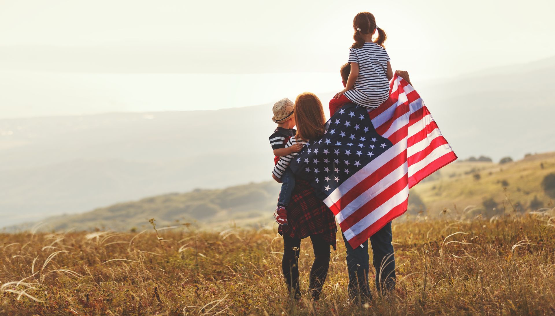 Family with American flag on a hilltop, child on shoulders, golden field and sky. | Bencomo Motorsport LLC