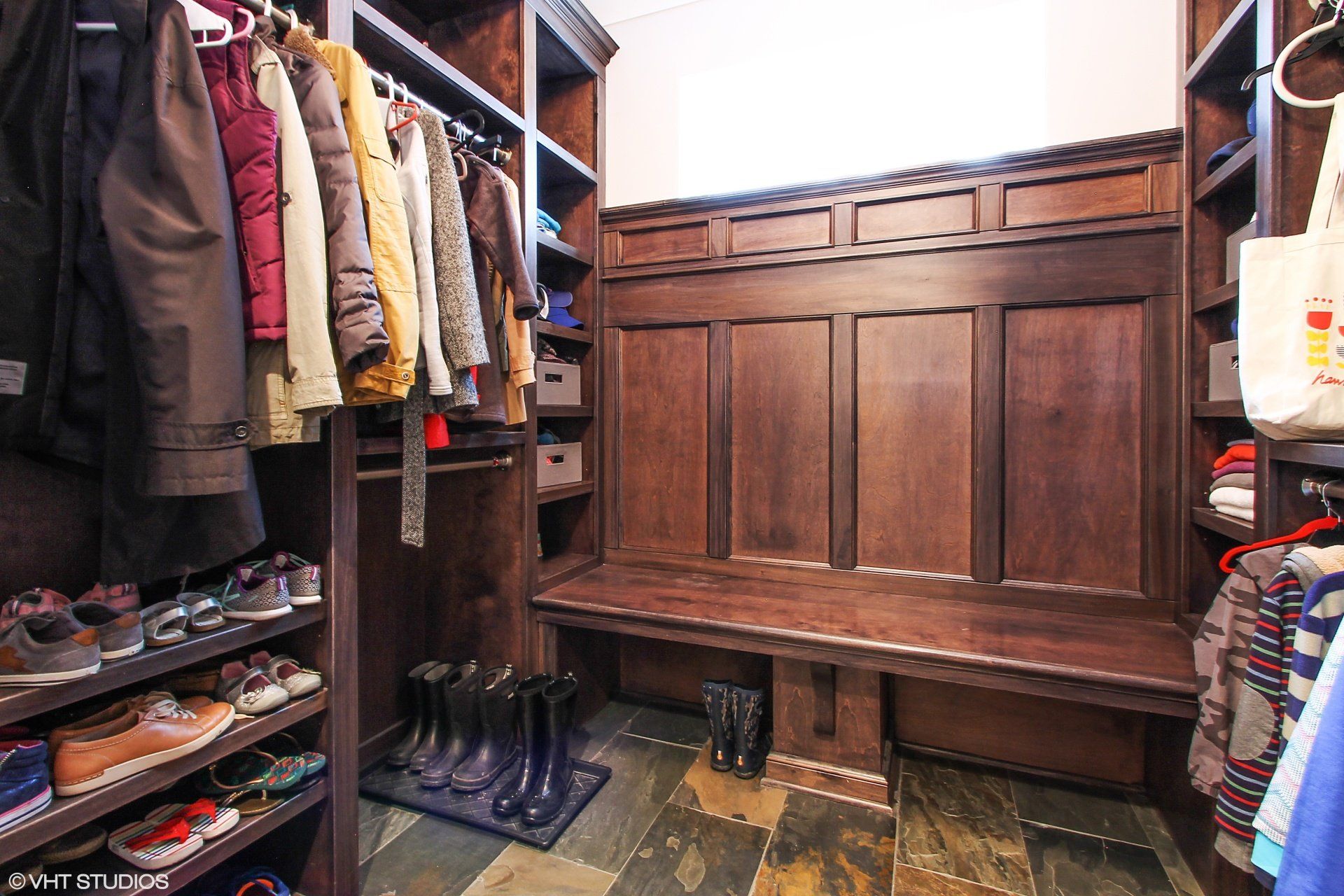 A walk in closet with a bench and shelves filled with clothes and shoes.