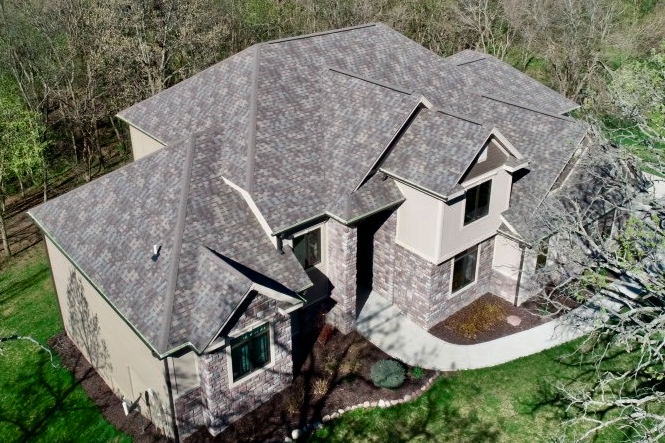 Aerial view of a house with a brown roof, tan siding, and stone accents, surrounded by green grass and trees.