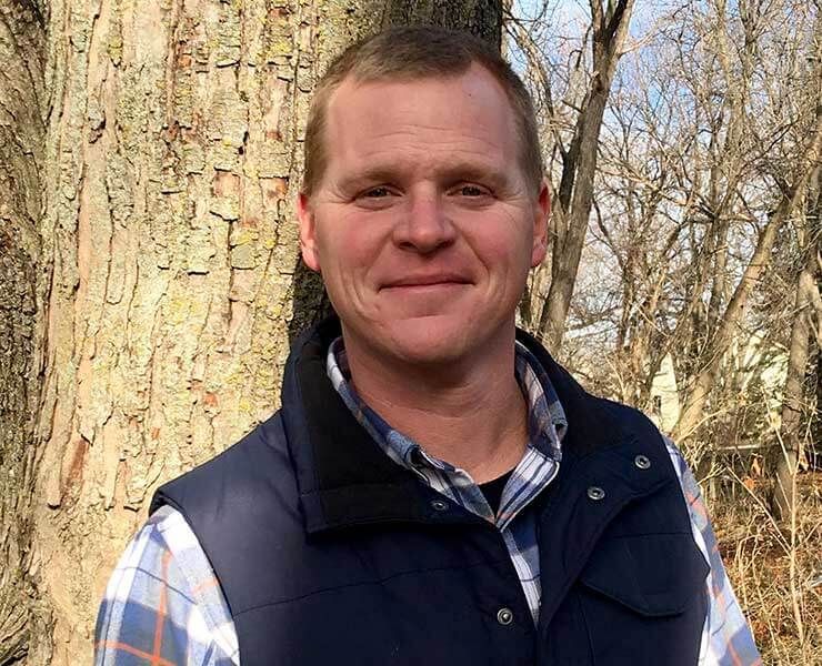 A man in a plaid shirt and vest is standing in front of a tree.