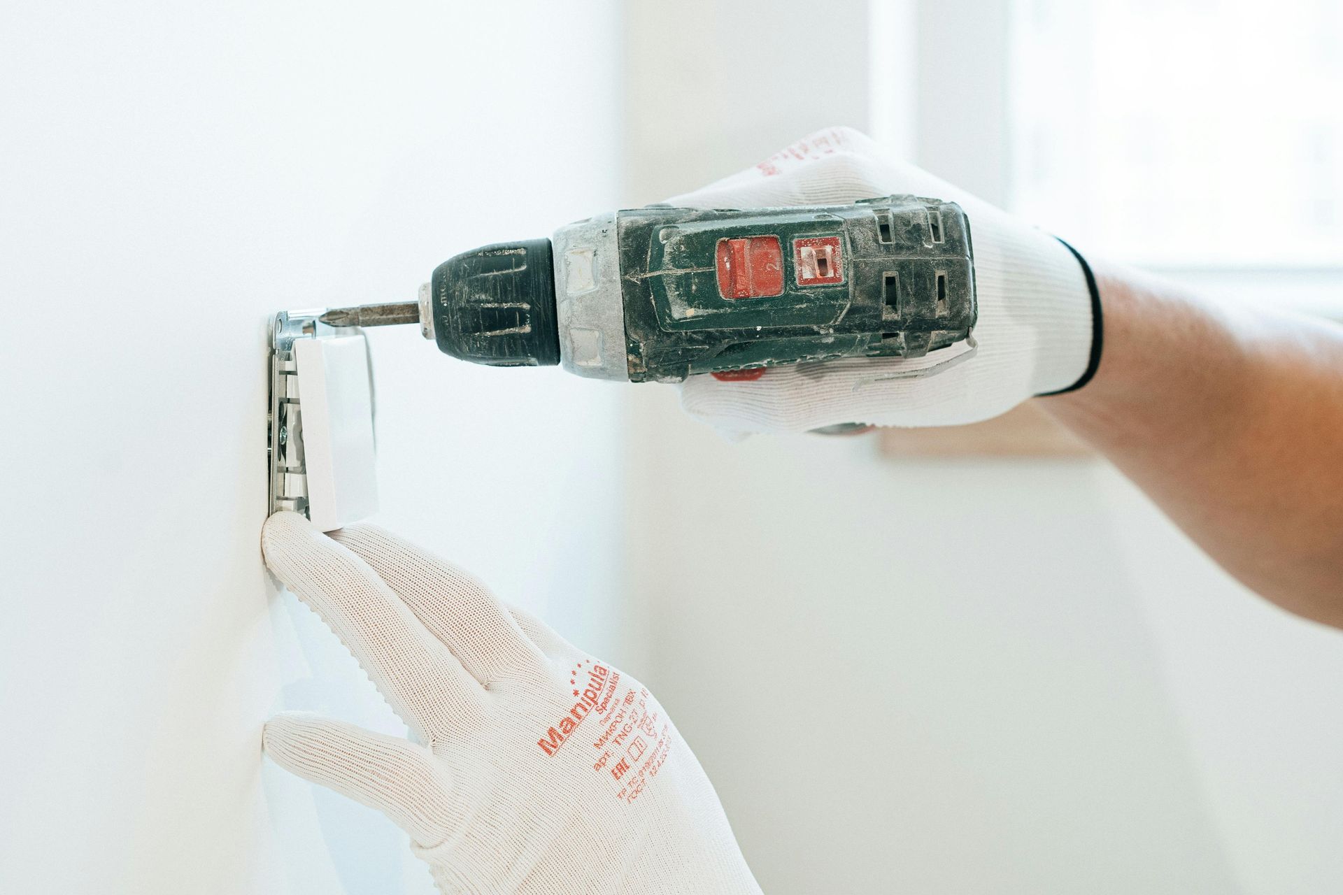 Person installing electrical outlet, using a drill. Hands wearing white gloves. White wall in the background.