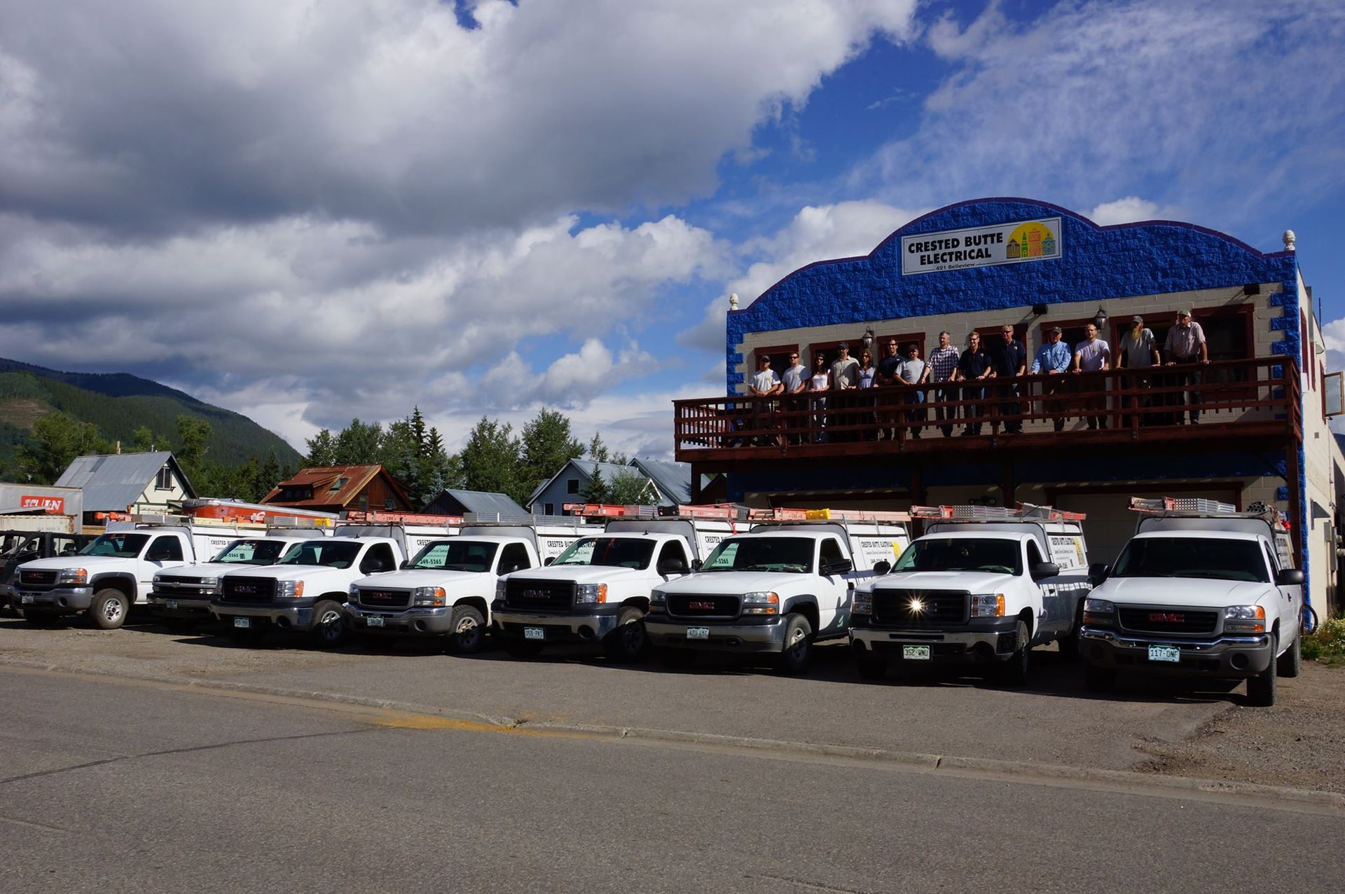 White trucks parked in front of a blue building with people on a balcony, mountain backdrop.