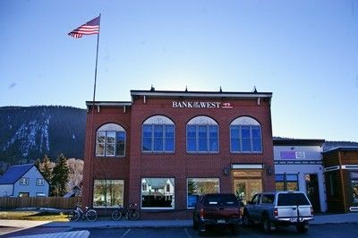 Bank building with American flag, parked cars. Red brick facade. Blue sky, mountain backdrop.
