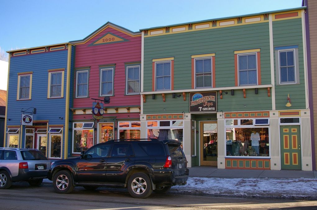 Colorful storefronts in a row, blue, red, and green, with cars parked in front, under a bright sky.